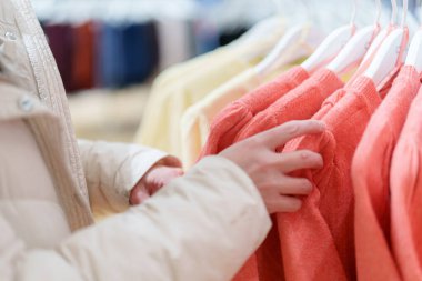 Bir insan giyim mağazasında askıda asılı renkli kazaklara bakıyor. Arka plandaki diğer pek çok giysiyle birlikte ortam parlak..