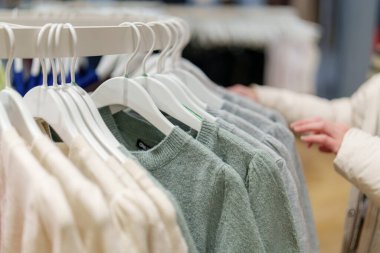 Alışverişçinin biri perakende mağazasının rafındaki kazaklara bakıyor. Etrafta başka renkler asılı dururken kişi gri bir kazağa dokunur. Mağazada bir sürü kıyafet seçeneği var..