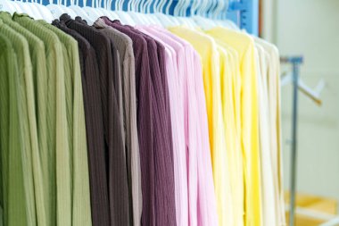 Perakende mağazasında bir sıra elbise askıda asılı duruyor. Giysilerde yeşil, kahverengi, pembe ve sarı olmak üzere farklı renkler gösteriliyor. Müşteriler seçenekler ve stiller arar.