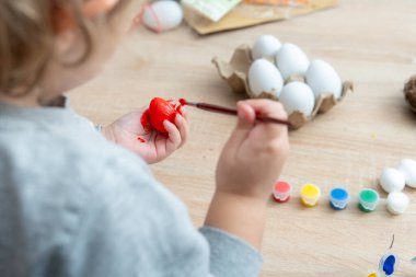 Küçük çocuk ahşap bir masanın üzerinde fırçayla kırmızı bir Paskalya yumurtası boyuyor. Masada boya, sade yumurta ve bayram için gerekli malzemeler de var..