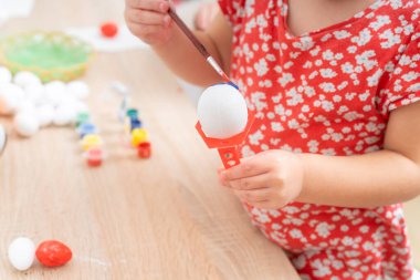 Genç bir kız beyaz bir Paskalya yumurtasını bir fırça ve mavi boyayla dikkatlice boyar. Masada çeşitli boyalar ve yumurtalar var. Bahar geleneği için hazırlar..