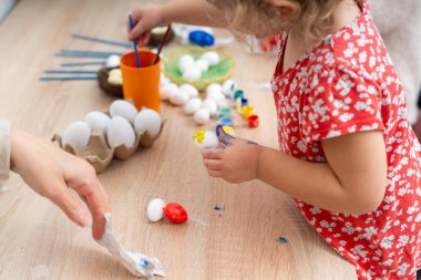 Kırmızı çiçekli bir elbise giyen genç bir kız ahşap bir masada Paskalya yumurtası boyuyor. Paskalya tatiline hazırlanırken annesi ona yardım ediyor..