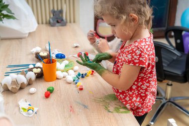 Masada yumurtaları süsleyen yeşil boyalı küçük bir çocuk görülüyor. Paskalya yumurtası dekorasyonu için boya, yuva ve diğer sanat malzemeleri masada..