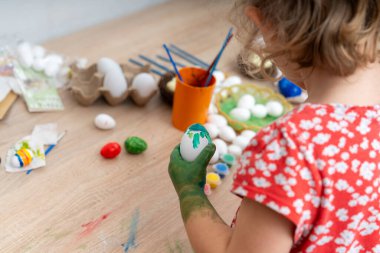 Kırmızı çiçekli gömlekli küçük bir kız Paskalya yumurtalarını, Paskalya bayramında boyalar, fırçalar ve yumurtalarla ahşap bir masaya boyayarak bayram sahnesi yaratıyor..