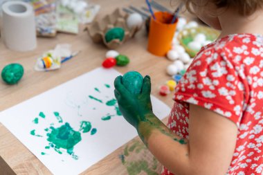 Yeşil boyalı elleri olan genç bir çocuk süslü bir Paskalya yumurtası tutuyor. Çocuk, resim malzemeleriyle kaplı bir masanın üzerine yeşil el izlerini de koydu..