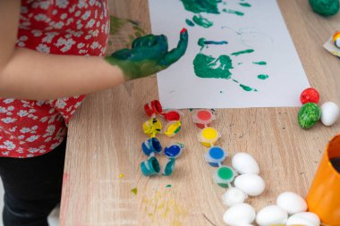 Ellerinde yeşil boya olan bir çocuk, paskalya yumurtalarının yanında kağıt üzerine boya ile resim yapıyor. Masa ahşap. Parlak, neşeli ve neşeli bir sahne..