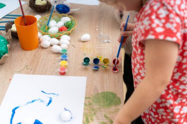 Genç bir kız, paskalya yumurtalarını rahat bir ev ortamında bir masada boya ve fırçayla süslüyor. Pastel renkler, bir sepet yumurta ve devam eden bir tablo var..