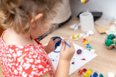 Küçük bir çocuk dikkatlice mavi boyalı bir Paskalya yumurtası boyuyor. Çocuğun elleri boyayla kaplı. Diğer boyanmış yumurtalar ve boya malzemeleri etraflarını sarar..
