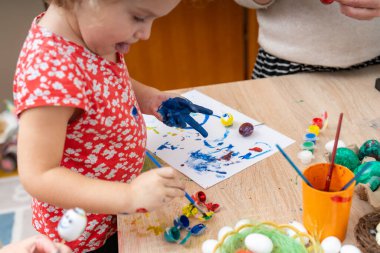 Kırmızı elbiseli küçük bir kız tahta bir masada paskalya yumurtası boyuyor. Etrafta boyalar, fırçalar ve yumurtalar var, neşeli bir Paskalya atmosferi yaratıyorlar..