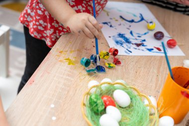Boyalı fırçalı bir çocuk renkli boyalarla kaplı bir masada paskalya yumurtası boyar. Masada yumurtalarla dolu küçük bir sepet var. Tatil için süslenmeye hazır..