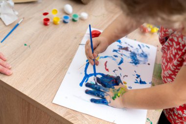 Genç bir çocuk mavi boya kullanarak beyaz kağıda resim yapıyor, yanında da renkli boyalar ve boş yumurtalar var. Etkinlik Paskalya tatili boyunca evde gerçekleşir..