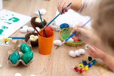 İki çocuk boya fırçasıyla Paskalya yumurtası boyuyor. Yumurtalar, boyalar ve yuvalar aile ile bahar tatili aktivitesi için masada..