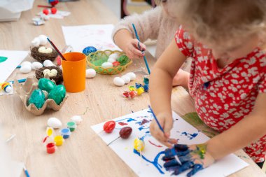 İki küçük çocuk Paskalya yumurtalarını boyuyor ve kağıt üzerinde renkli sanat eserleri yaratıyorlar. Masadaki boya, fırça ve yumurtalarla bahar tatiline hazırlanıyorlar..
