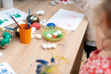 Paskalya yumurtalarını evde boyayan bir insan, onları yuvalara ve bir sepete yerleştiriyor. Masanın üzerinde el izi ve boya olan bazı bitmiş sanat eserleri de var..