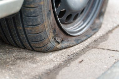 Patlak lastikli bir araba caddede duruyor. Lastik yıpranma ve hasar gösteriyor. Asfalt ıslak ve çatlakları gösteriyor. Bu sahne, gündüz vakti meydana gelen yaygın bir yol kenarı sorununu gösteriyor..