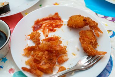 A plate shows leftover shrimp pieces after a meal. One shrimp is intact while the others are eaten. A fork rests nearby. The setting is casual and colorful, suggesting a joyful meal.