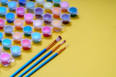 Parlak sarı bir yüzeye yerleştirilmiş çeşitli renklerde küçük boya bardakları koleksiyonu. Yakınlarda boyama faaliyetlerine hazır üç fırça var..