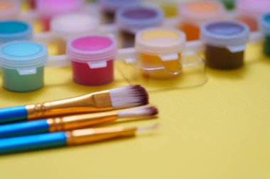 Üç boya fırçası, çeşitli renklerde küçük boya kaplarının yanında sarı bir yüzeyde duruyor. Stüdyo, düzenli ve kullanıma hazır sanat malzemeleriyle dolu..