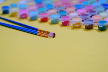 İki boya fırçası çeşitli küçük boya kaplarının yanında sarı bir yüzeyde duruyor. Çalışma alanı parlak ve düzenli görünüyor. Yaratıcı çalışmaya hazır..