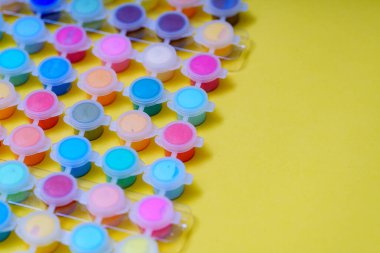 Farklı renklerle dolu küçük bardaklar sarı bir yüzeyde bir ızgaraya yerleştirilir. Boya bardakları yaratıcı projeler için çeşitli parlak gölgeler sergiliyor.