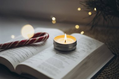 Açık İncil, yanan mum ve evdeki pencere eşiğinde Noel şekeri. İncil ve Bokeh ışıklarıyla samimi bir Noel arkaplanı.