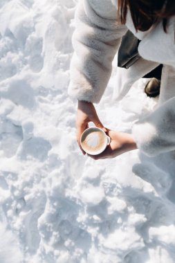 Bir kadının elinde bir bardak kapuçino, karla kaplı bir arka plan, üst manzara. Bir kadın kışın dışarıda kahve içer..