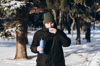 Termoslu genç sakallı bir adam kış ormanında sıcak bir içkinin tadını çıkarıyor. Parkta yürürken bir adam çay içer. Kış ormanındaki bir termostan çay..