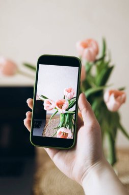 Bir kadın, ahşap bir masadaki dizüstü bilgisayarın yanında vazoda lalelerin fotoğrafını gösteren bir akıllı telefonu tutuyor. Konforlu çalışma alanı teknoloji ve doğal güzelliği harmanlıyor, yaratıcılık ve verimliliğe ilham veriyor..
