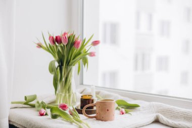 Ev penceresinin kenarında bir kahve fincanı, mum ve pembe lalelerin yakın çekimi. Doğal ışıklandırması olan rahat ve kadınsı bir sahne, yaşam tarzı, kendine bakım, ev dekorasyonu ve rahatlama içeriği için mükemmel..