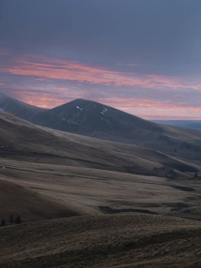 A wide-angle scenic view of dark, rolling hills under a deep purple sky with glowing orange light on the horizon.