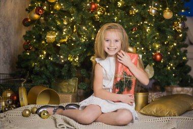 Mutlu şirin kız, kırmızı hediye kutusuyla Noel ağacının yanında oturuyor..