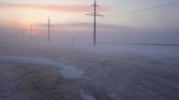 Nieve en el camino helado, el sol en la niebla. Los postes de energía alinean la carretera. Nieve barre la carretera en una helada severa al atardecer — Vídeo de stock