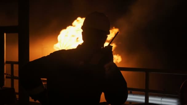 Silhouet van een man tegen een vlam. Een man in een helm op een achtergrond van vuur. Brandweerman silhouet met een walkietalkie tegen de achtergrond van vlammen. Goedenacht. Man kijkt naar vuur, uitzicht van achteren. — Stockvideo