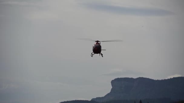 The helicopter is flying towards us. Orange helicopter. Flies different sides forward. Against the background of mountains and sky — ストック動画