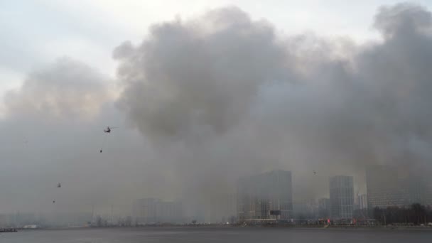 Helicopters fly with bambi bucket. Rescue helicopters over river. City is in smoke. Fire. Houses on embankment in smoke — Vídeo de Stock