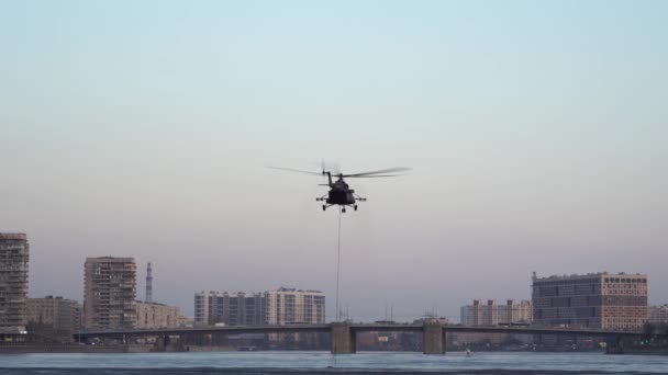 ヘリコプターは川でバンビバケツを拾います。ホバー。後ろの景色。後ろの川に架かってる。ヘリコプターはバケツで離陸する。サンクトペテルブルク, — ストック動画