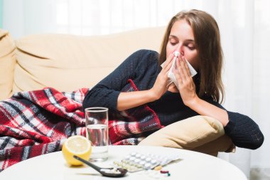 Sick woman lying on sofa under wool blanket sneezing and wiping nose clipart