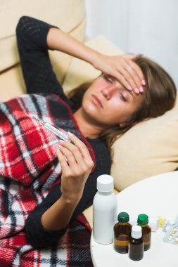 Yün battaniyenin altında kanepeye yatan ve termometreye bakan hasta kadın