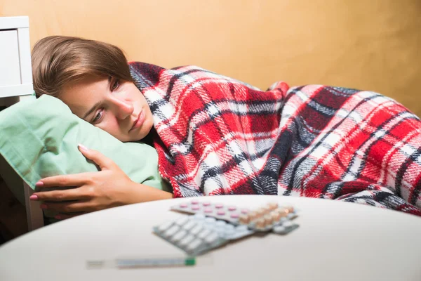 Mujer enferma acostada en la cama bajo una manta de lana mirando el termómetro y las pastillas — Foto de Stock