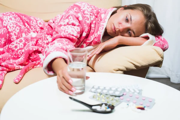 Больная женщина лежит в постели в халате и смотрит на термометр и таблетки. — стоковое фото