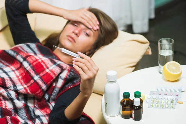 Wanita sakit berbaring di sofa di bawah selimut wol dan melihat termometer — Stok Foto