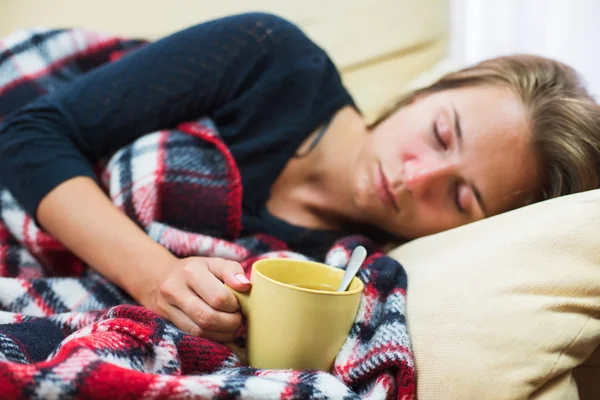 Mujer enferma acostada en un sofá debajo de una manta de lana con una taza de té — Foto de Stock