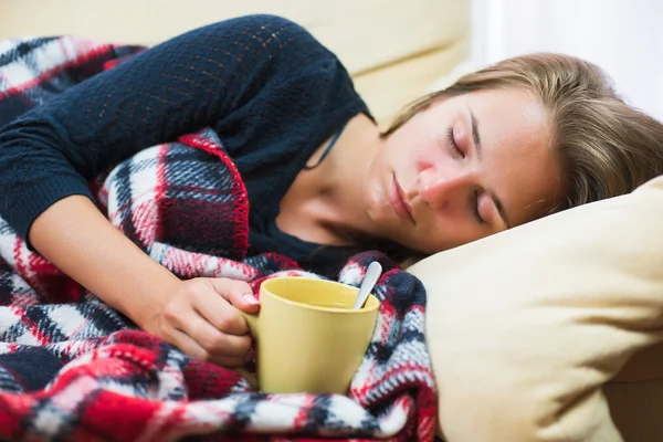 Mujer enferma acostada en un sofá debajo de una manta de lana con una taza de té — Foto de Stock