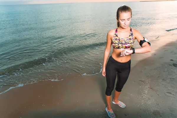Joven chica en forma mirando el reloj durante los ejercicios de la mañana en la playa amanecer — Foto de Stock