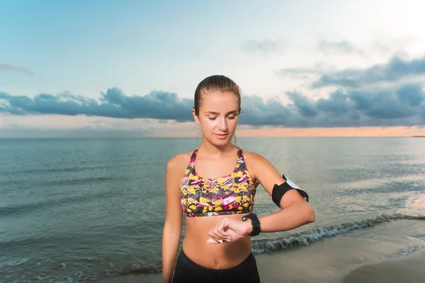 Chica en forma joven mirando el reloj durante los ejercicios de la mañana en la playa — Foto de Stock