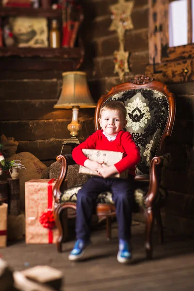 Small boy sitting in chair with present next to Christmas tree and gifts — Stock Photo, Image