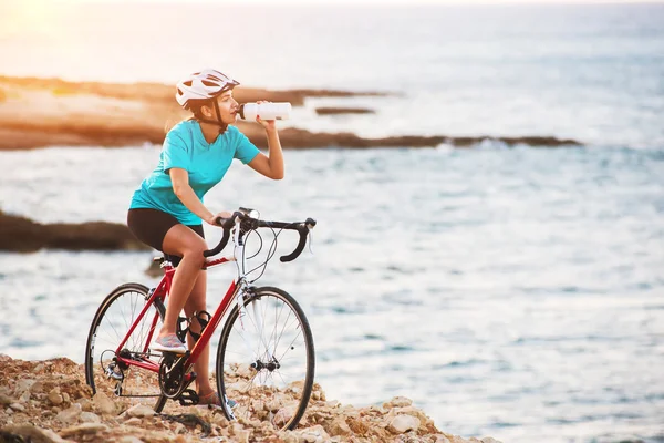 Ciclista feminina em pé em uma rocha com bicicleta e água potável — Fotografia de Stock