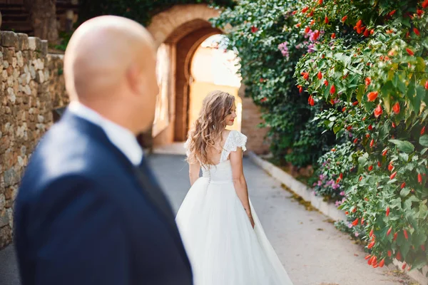 新郎は城のアーチの背景に立って白いドレスで彼の美しい花嫁を見たり賞賛します 屋外結婚式 — ストック写真