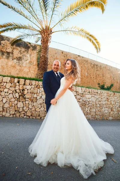 Newlyweds hug and smile, at the entrance to the castle against the background of a stone wall and palm trees