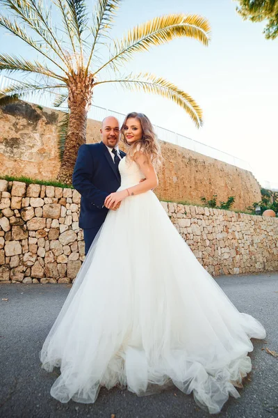 Newlyweds hug and smile, at the entrance to the castle against the background of a stone wall and palm trees